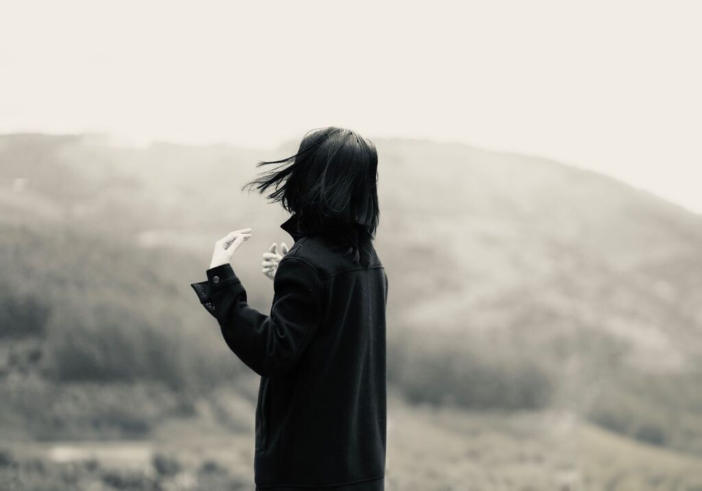 A woman stands with her back turned, looking towards a misty and hilly landscape, creating a sense of solitude and contemplation.